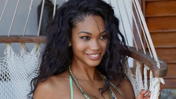 Chanel Iman poses in front of a hanging hammock chair in a green bikini and smiles off into the distance.